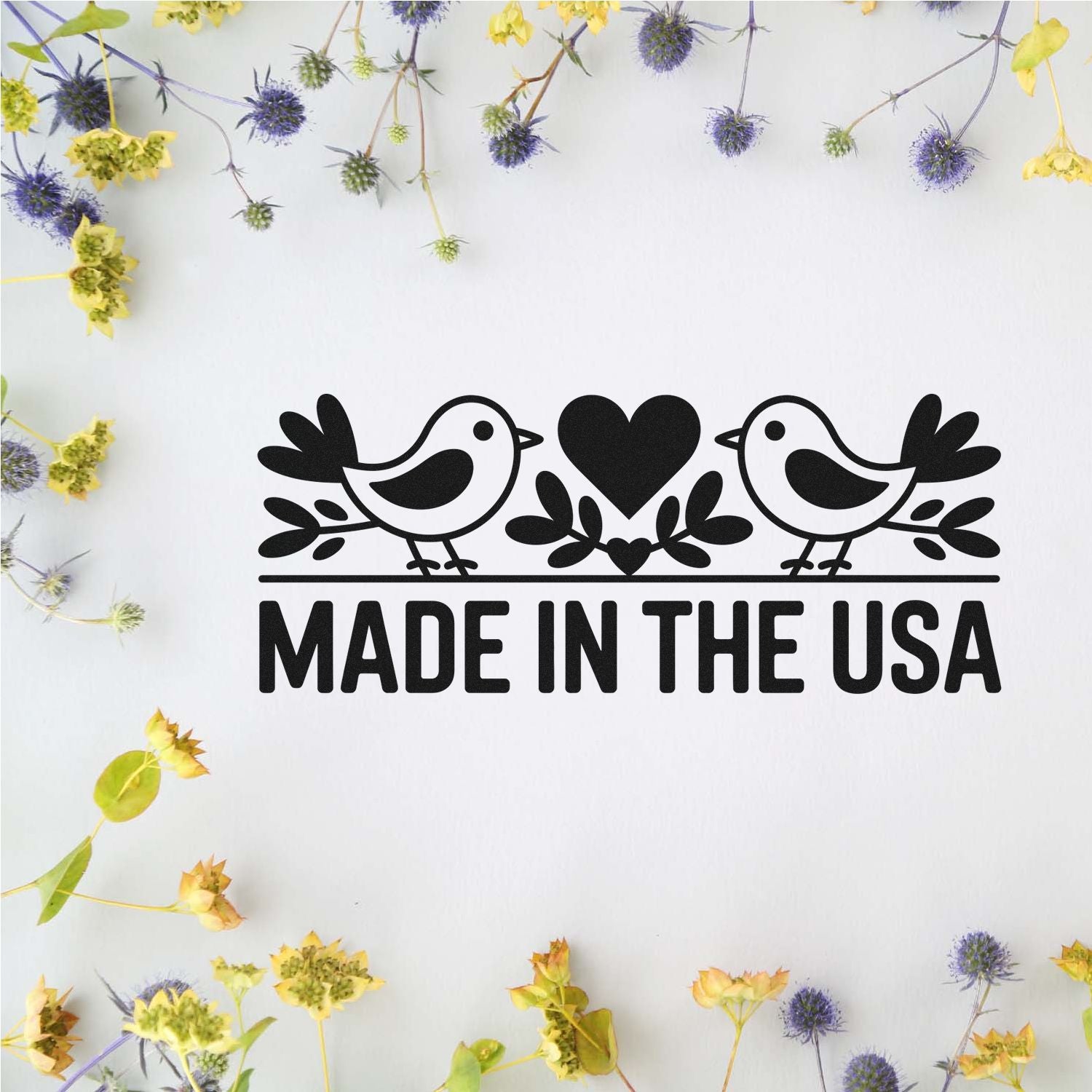 Black Made in the USA stamp seal with two birds, heart, and white background framed by yellow and purple dried flowers.