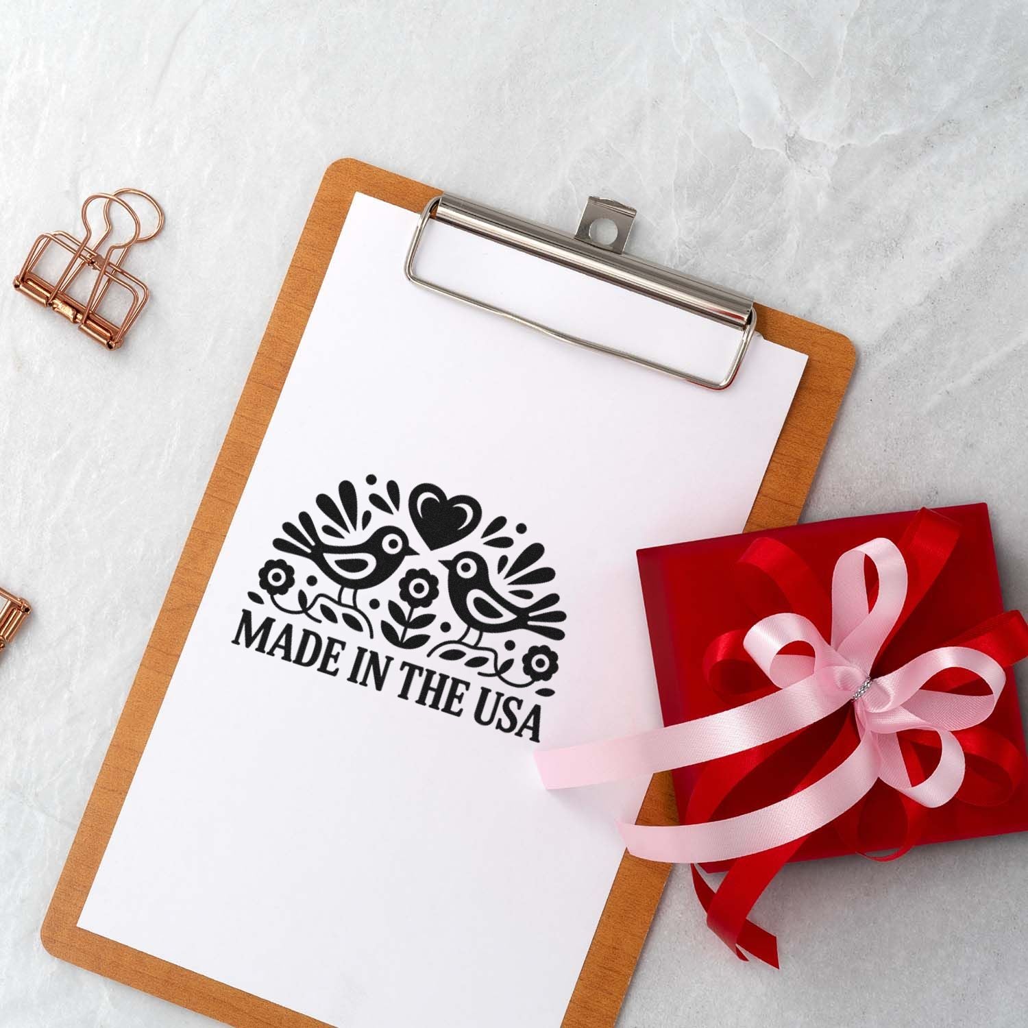 White clipboard displays a black Made in the USA stamp seal with birds and heart, beside a red gift box with pink ribbon.
