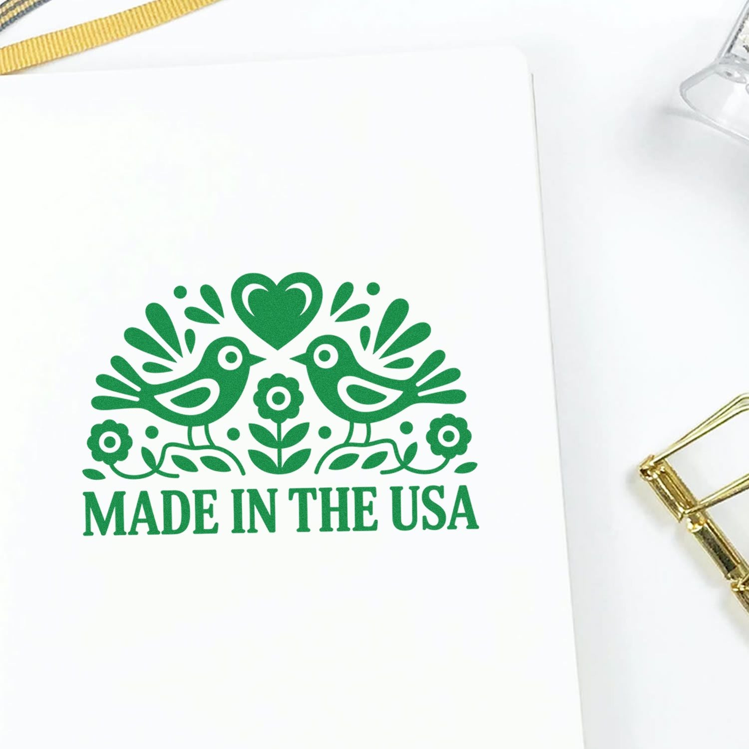 Green Made in the USA stamp seal with birds, heart and flowers on white paper, beside a gold clip.