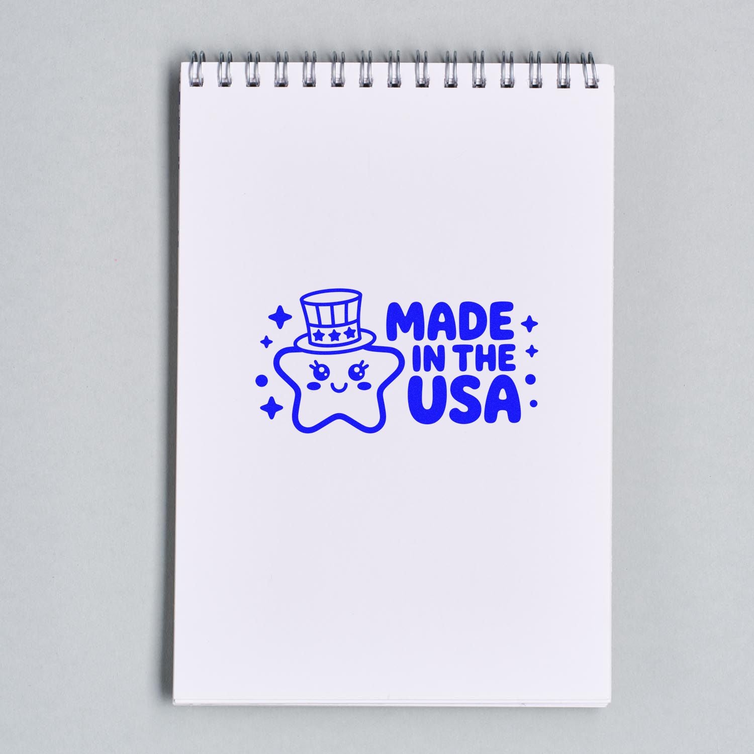 Blue Made in the USA rubber stamp featuring a smiling star in a top hat on a white spiral notebook.