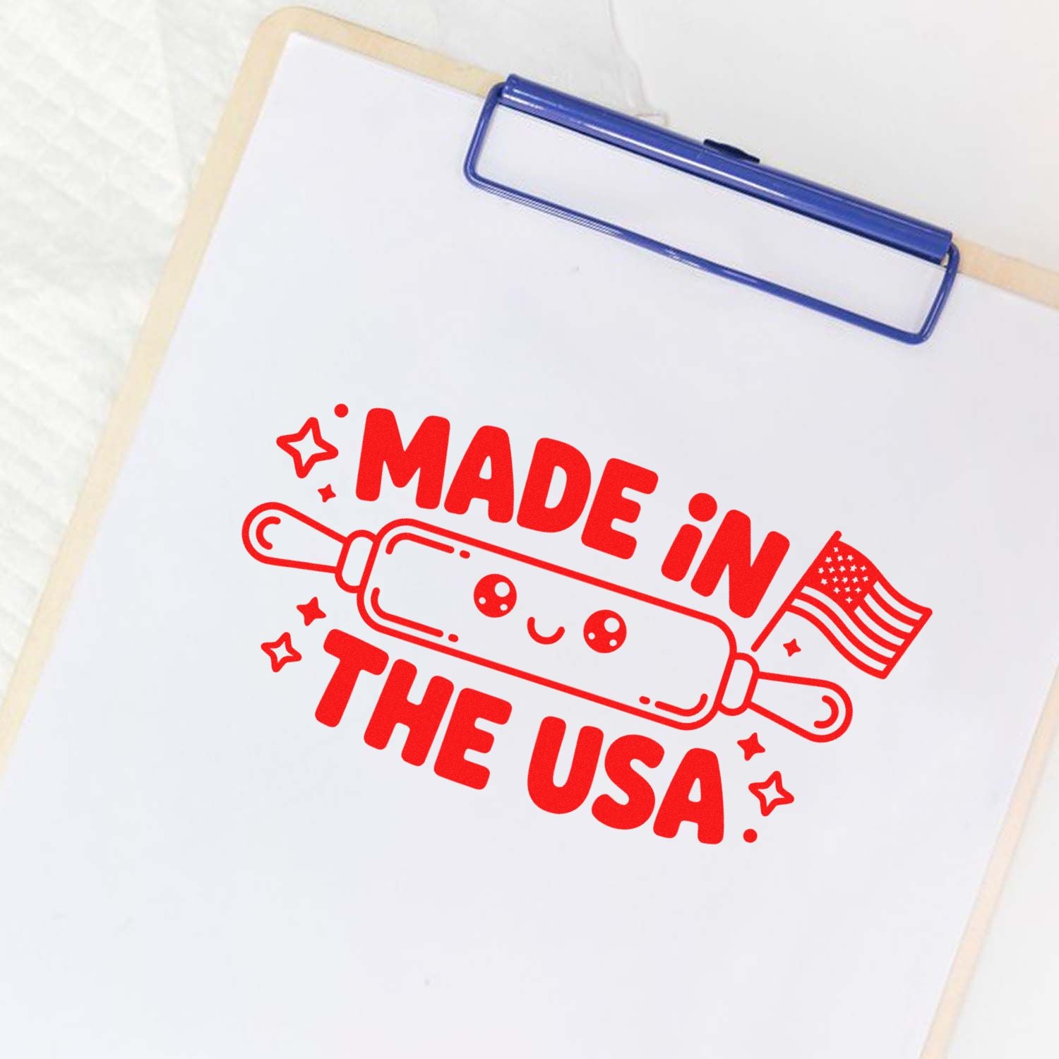 Red rubber stamp/seal design reading Made in the USA, with rolling pin, stars, and flag on white paper with blue clipboard clip.