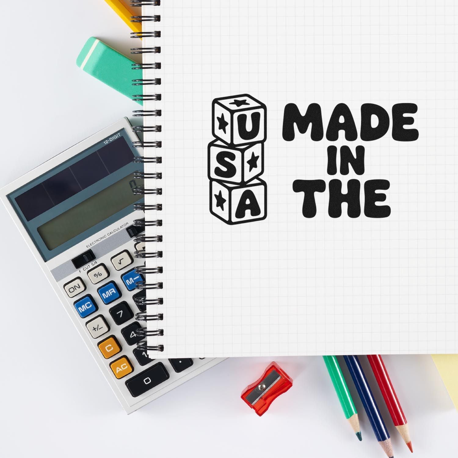 Black MADE IN THE USA stamp and seal on graph notebook beside calculator, colored pencils, and sharpener.