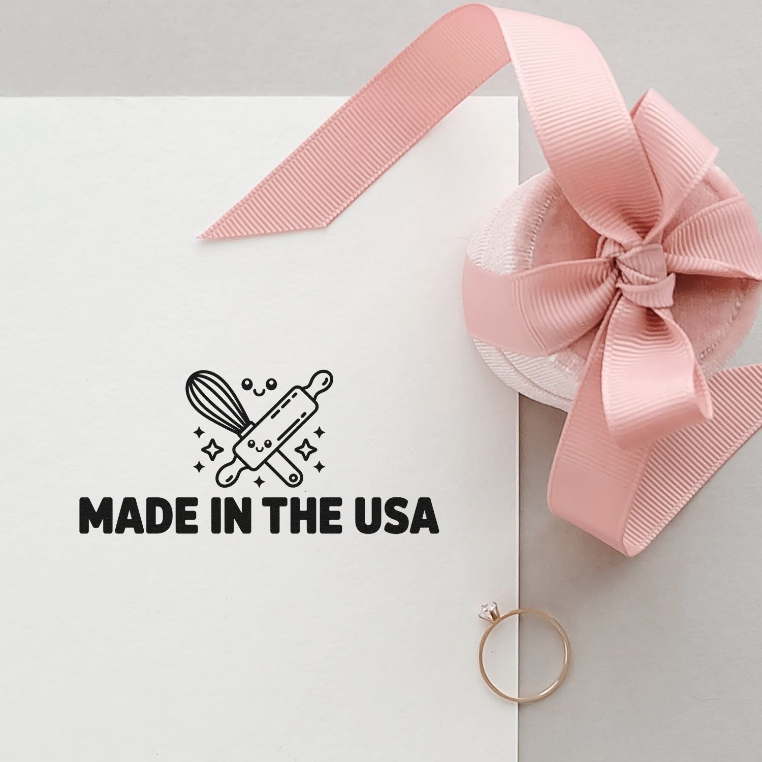 Pink ribbon-wrapped wax seal stamp package with "Made in the USA" logo on white, plus a gold ring accent.