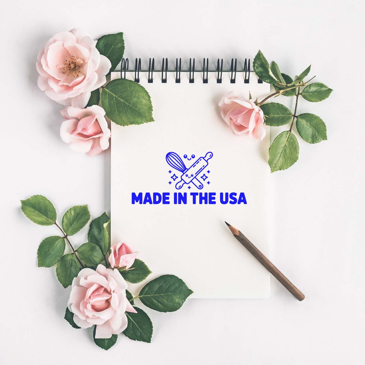 White spiral notebook with blue Made in the USA stamp logo, pink roses, green leaves, and a brown pencil.