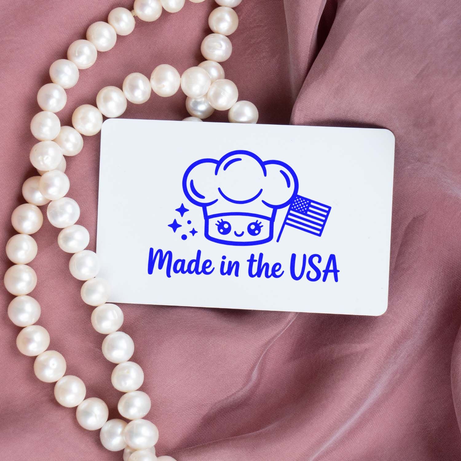 White stamp and seal card with blue Made in the USA chef icon and flag, on pink fabric with pearl beads