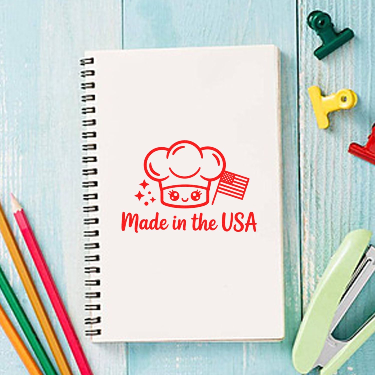 Red chef stamp seal reading Made in the USA on white spiral notebook, with colorful clips and pencils on blue wood.