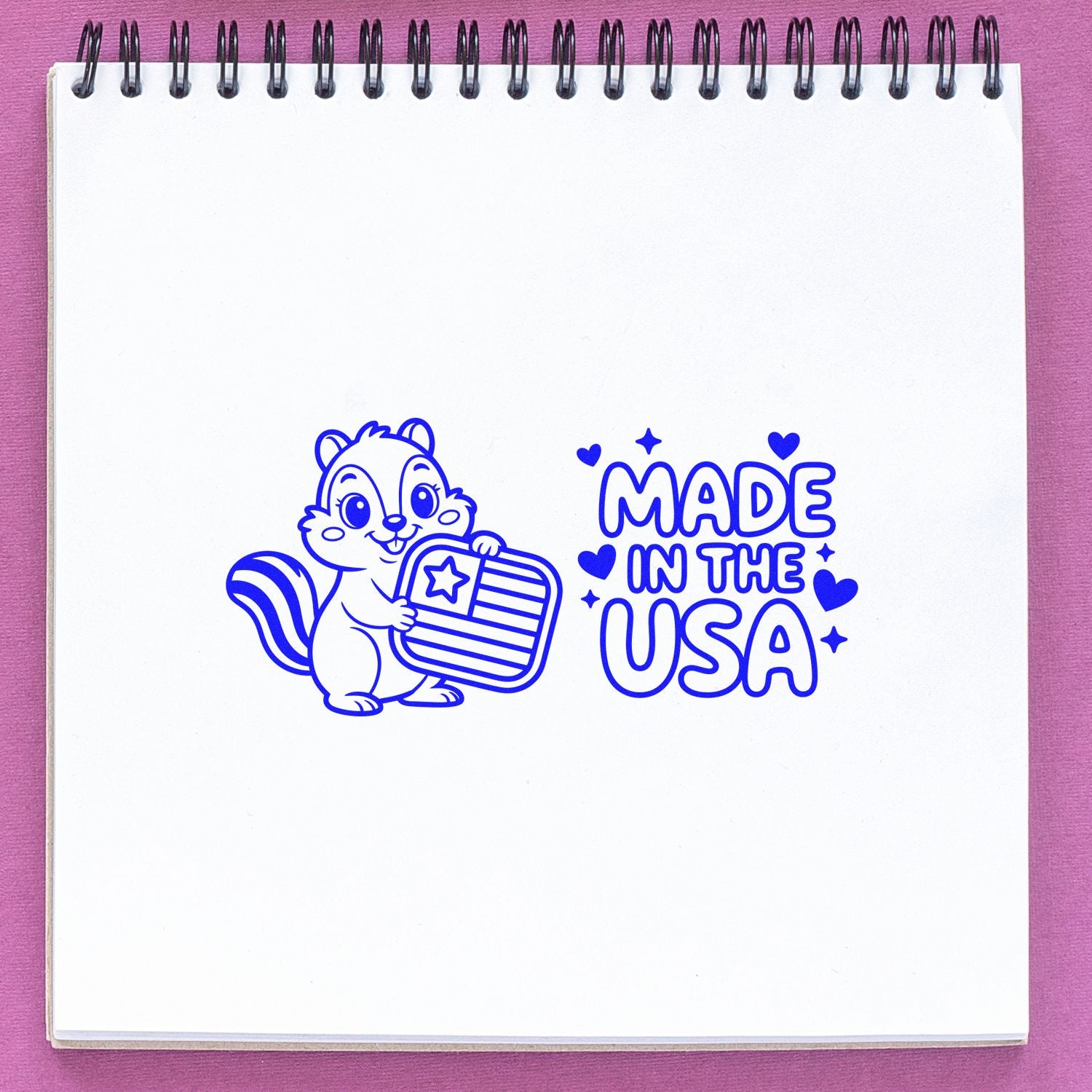 Blue patriotic stamp design with squirrel holding U.S. flag and Made in the USA text on white spiral notebook, pink background.