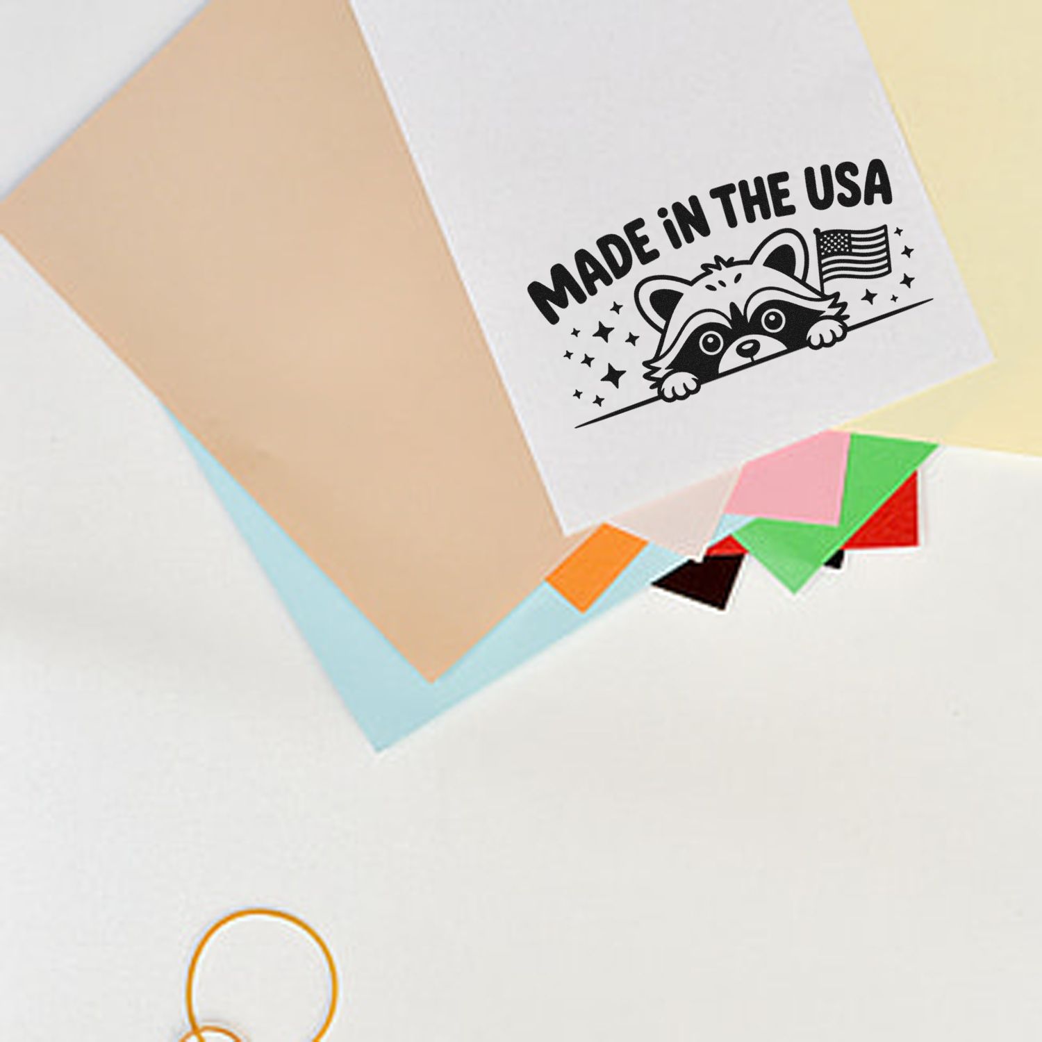 Black Made in the USA raccoon stamp with U.S. flag on white paper, surrounded by pastel seal sample sheets.