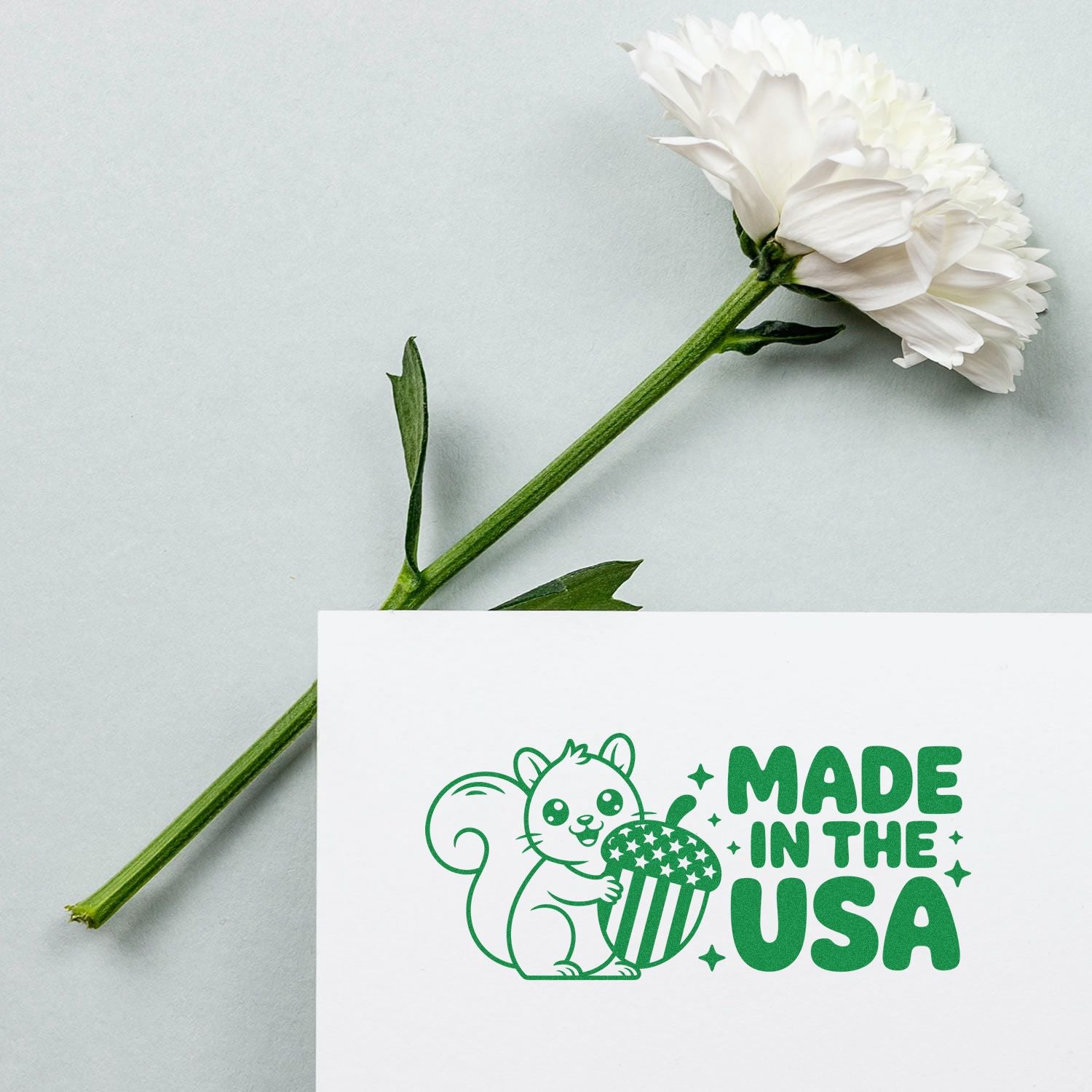 Green Made in the USA stamp/seal with squirrel and acorn on white card beside a white flower with green stem.