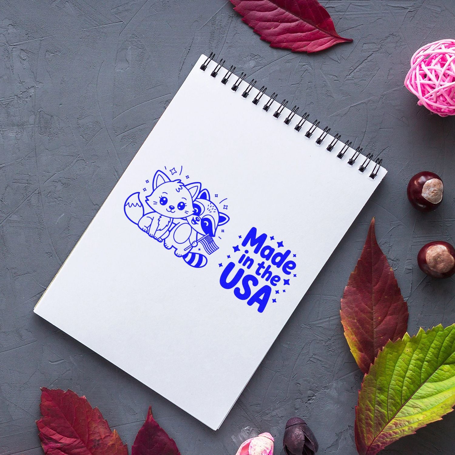 White spiral notebook with blue fox stamp and Made in the USA seal, on gray textured background with autumn leaves.