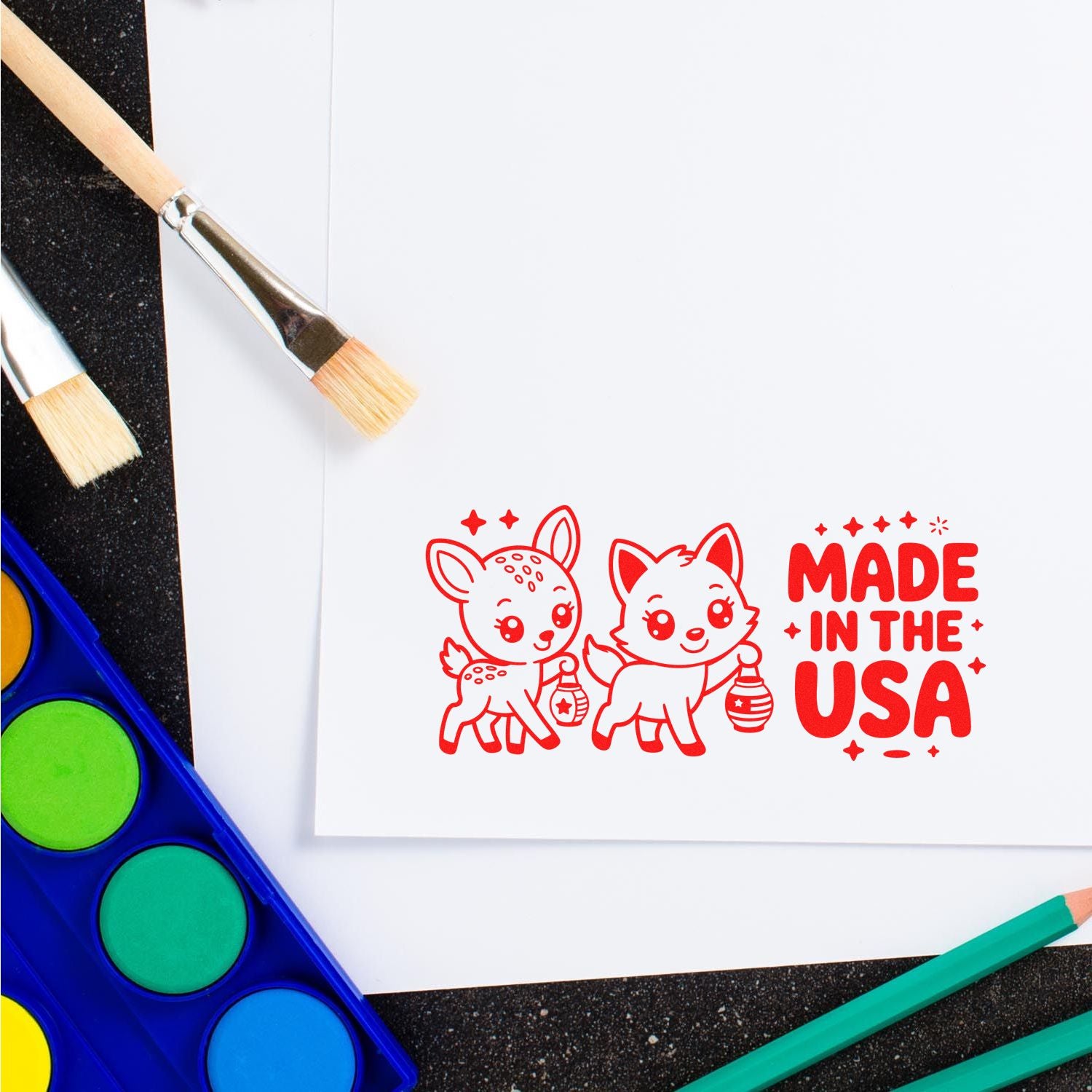 Red Made in the USA stamp seal with cartoon deer and fox on white paper, surrounded by paint brushes and bright watercolor palette