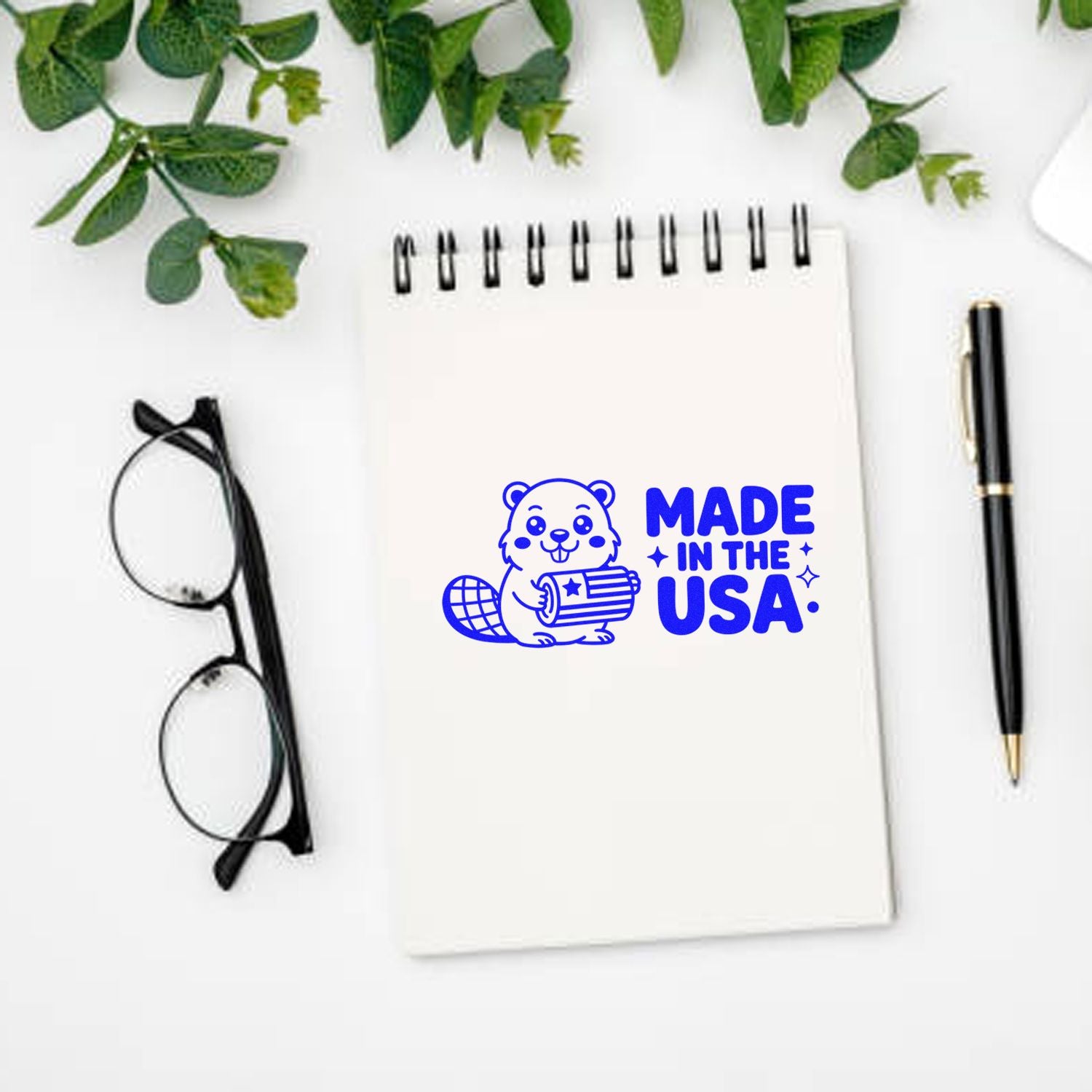 Blue Made in the USA stamp seal with beaver on white spiral notebook, beside black pen and glasses.