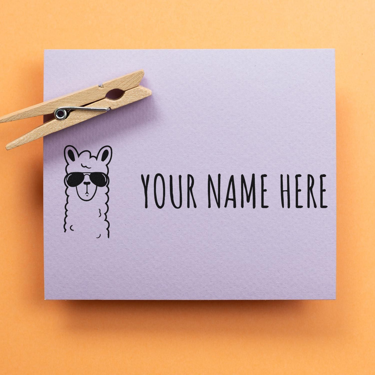 Lavender custom stamp-and-seal sample with black llama in sunglasses, YOUR NAME HERE text, and wooden clothespin on orange background.