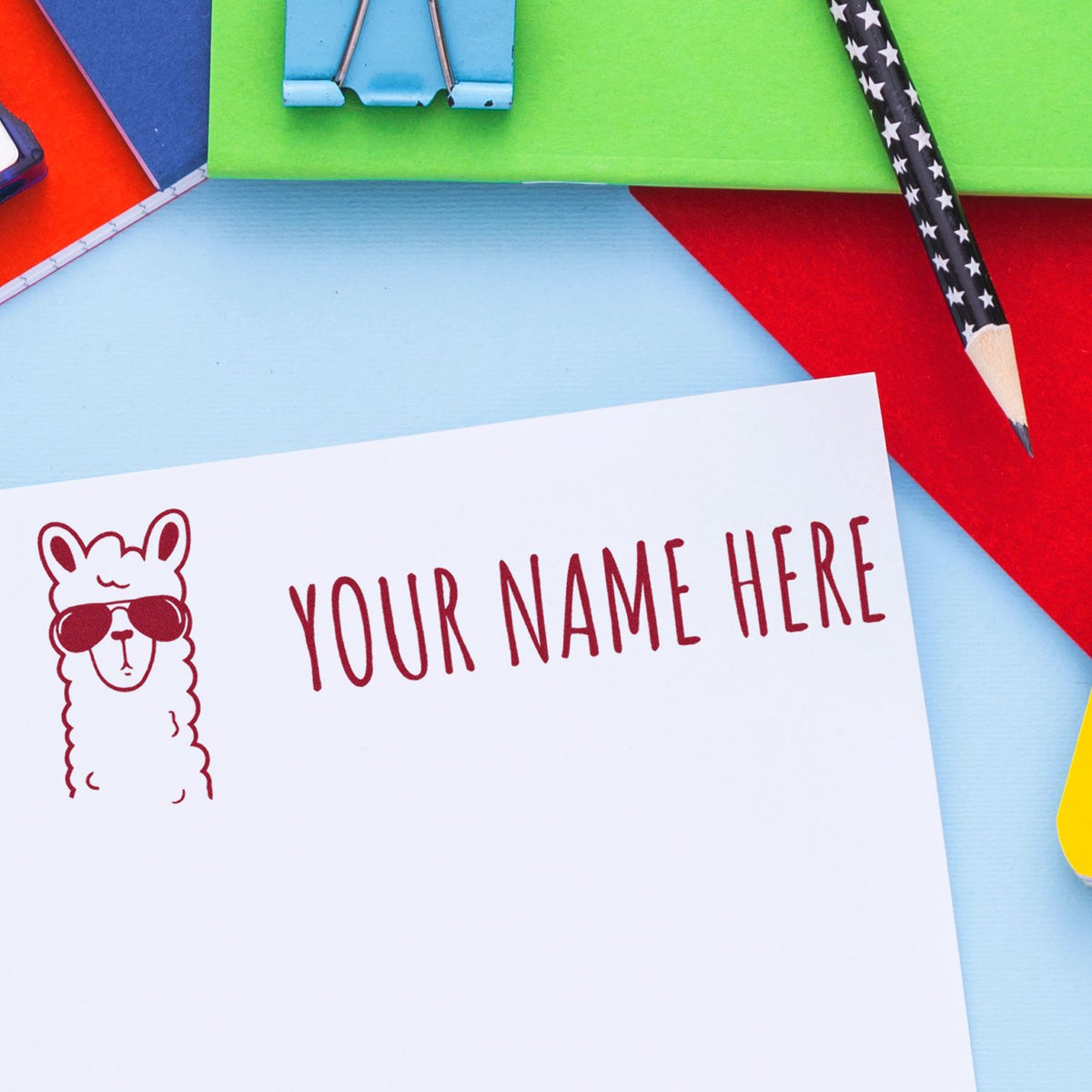 Red custom stamp seal with llama in sunglasses on a white card, surrounded by colorful stationery and pencils.