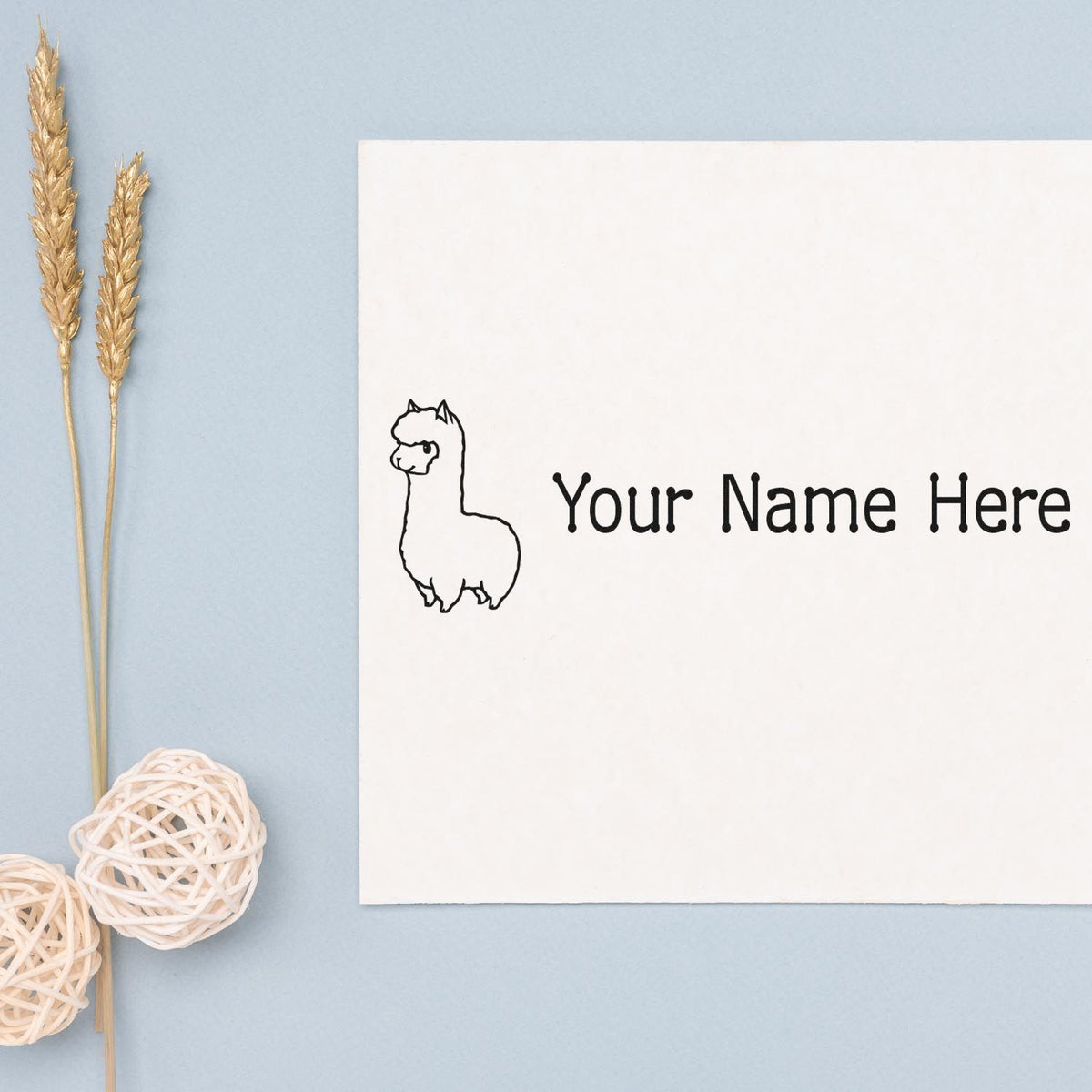 Custom llama stamp and seal on a white card with black text, set against a pale blue background with gold wheat and white wicker decor.