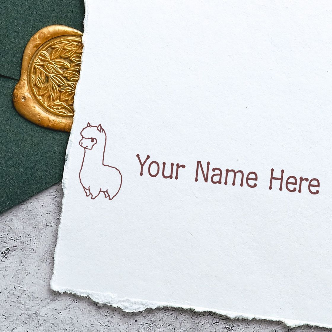 Personalized brown llama name stamp on white textured paper with a gold embossed wax seal on dark green fabric.