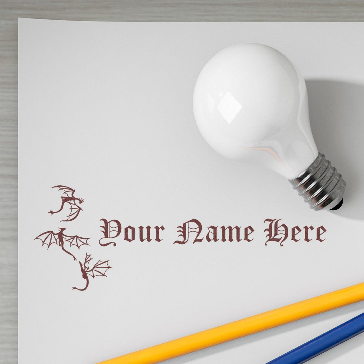 Custom stamp and seal with maroon dragon graphics and Your Name Here text on white paper beside a white lightbulb and pencils.