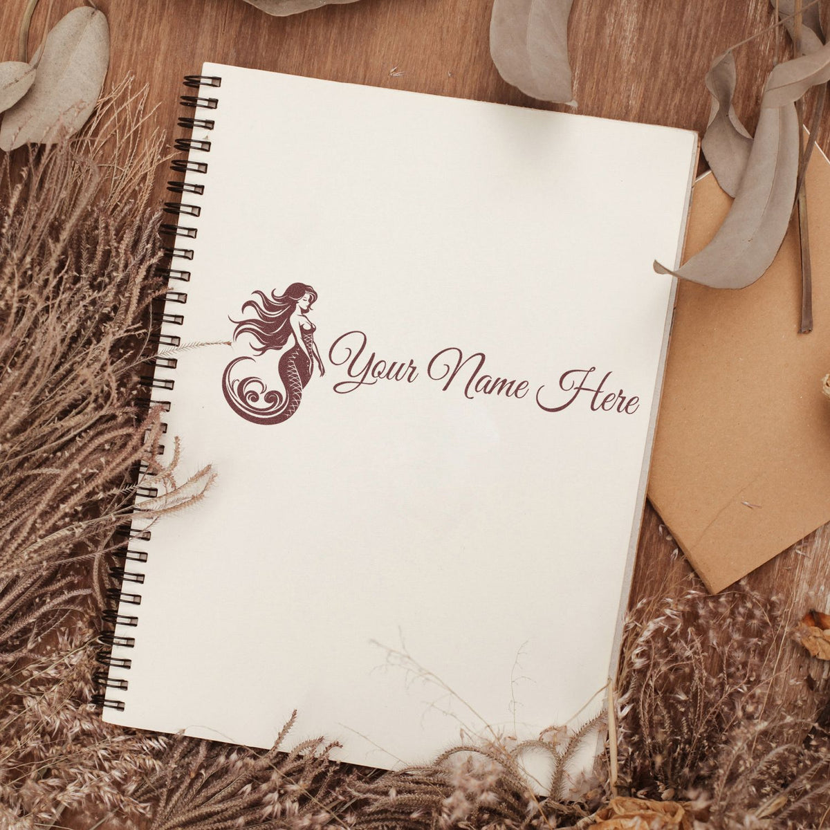 Maroon mermaid stamp and seal on a cream spiral notebook, surrounded by dried beige foliage on a warm wood background.