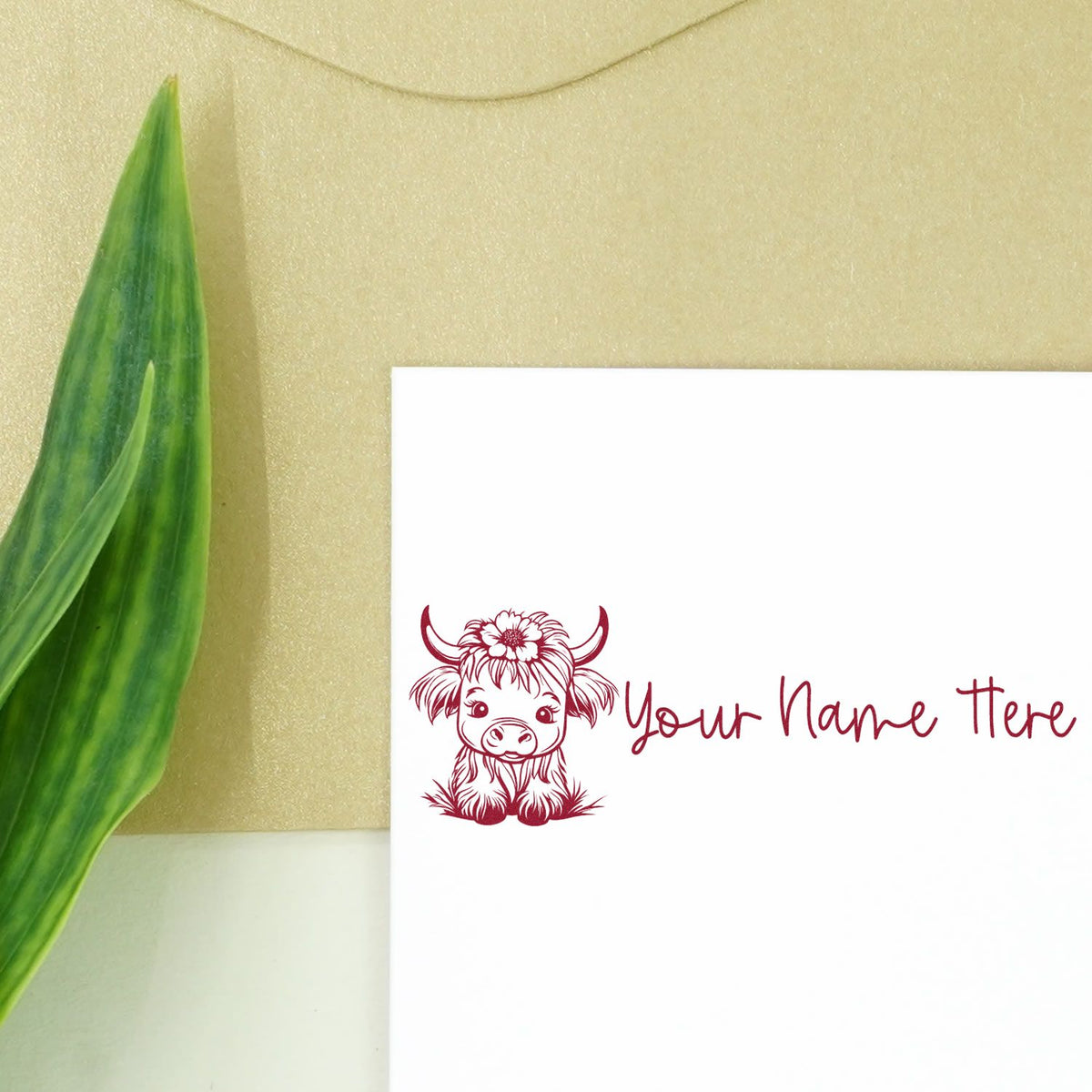 Custom maroon cow stamp seal with flower and Your Name Here on a white card, beside a green leaf on beige background