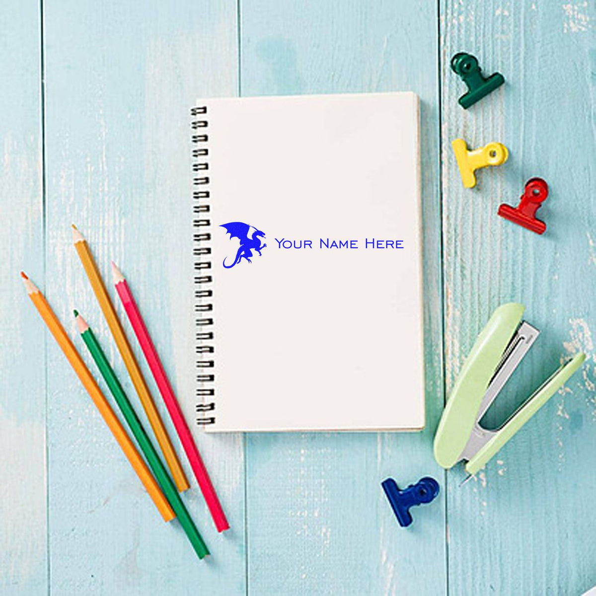 Custom blue dragon stamp and seal on white spiral notebook, with colorful pencils and binder clips on a light blue wood background.