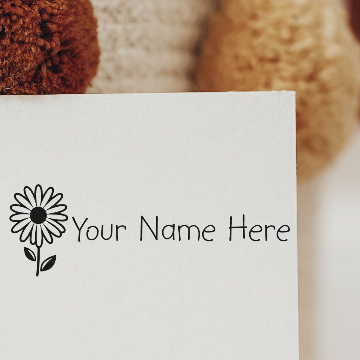 Black daisy logo stamp and seal with Your Name Here on white paper, against warm beige and brown background.
