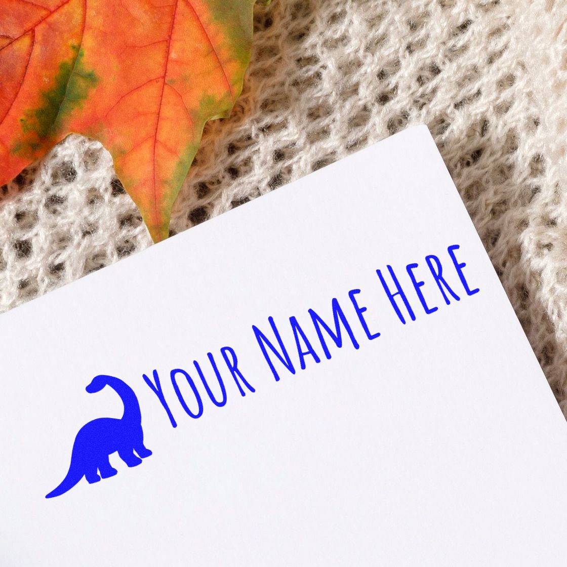 Custom blue dinosaur name stamp and seal on white paper, with YOUR NAME HERE, on beige knit fabric and orange leaf.