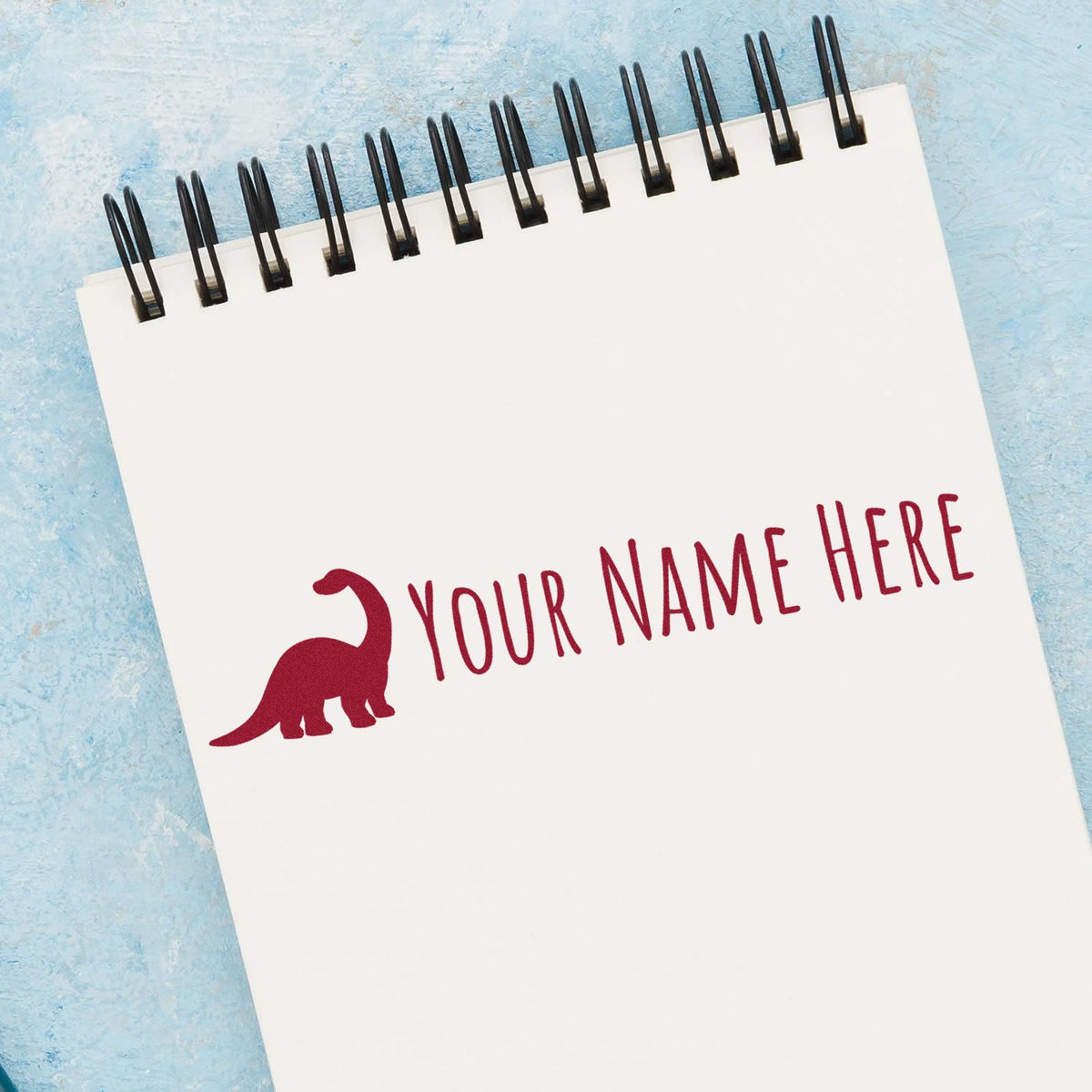 White spiral notebook featuring a red dinosaur name stamp, black spiral binding, on a blue textured background.