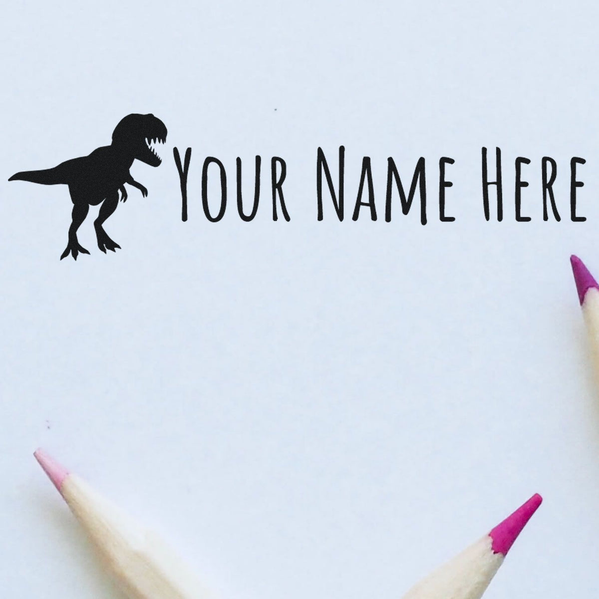 Black dinosaur custom name stamp with Your Name Here on white paper, framed by pink pencil tips.