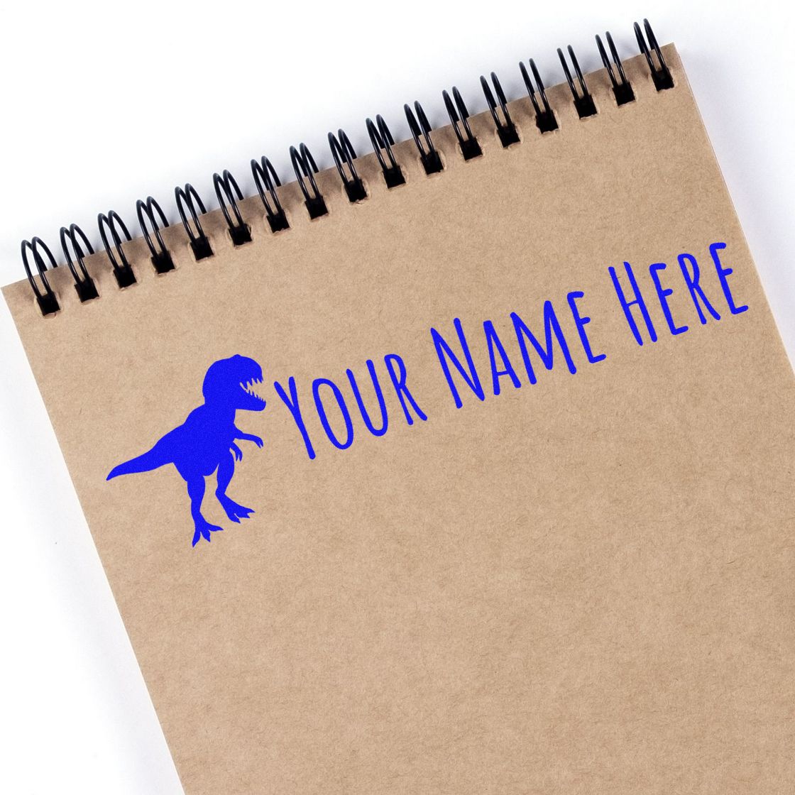 Kraft spiral notebook with bright blue dinosaur stamp and YOUR NAME HERE custom seal on tan paper.