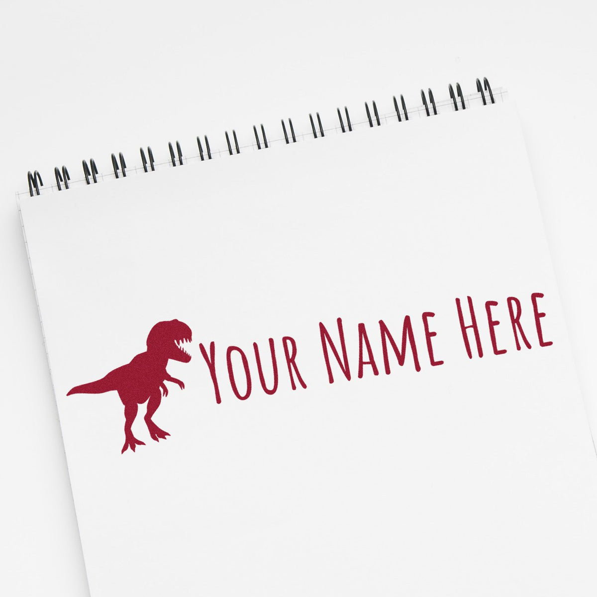 White spiral notebook with red custom stamp seal, dinosaur silhouette, and YOUR NAME HERE text.