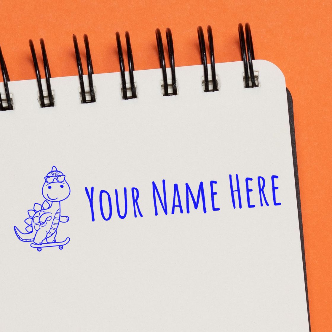 Blue custom stamp seal with dinosaur on skateboard and YOUR NAME HERE on a white spiral notebook, orange background.