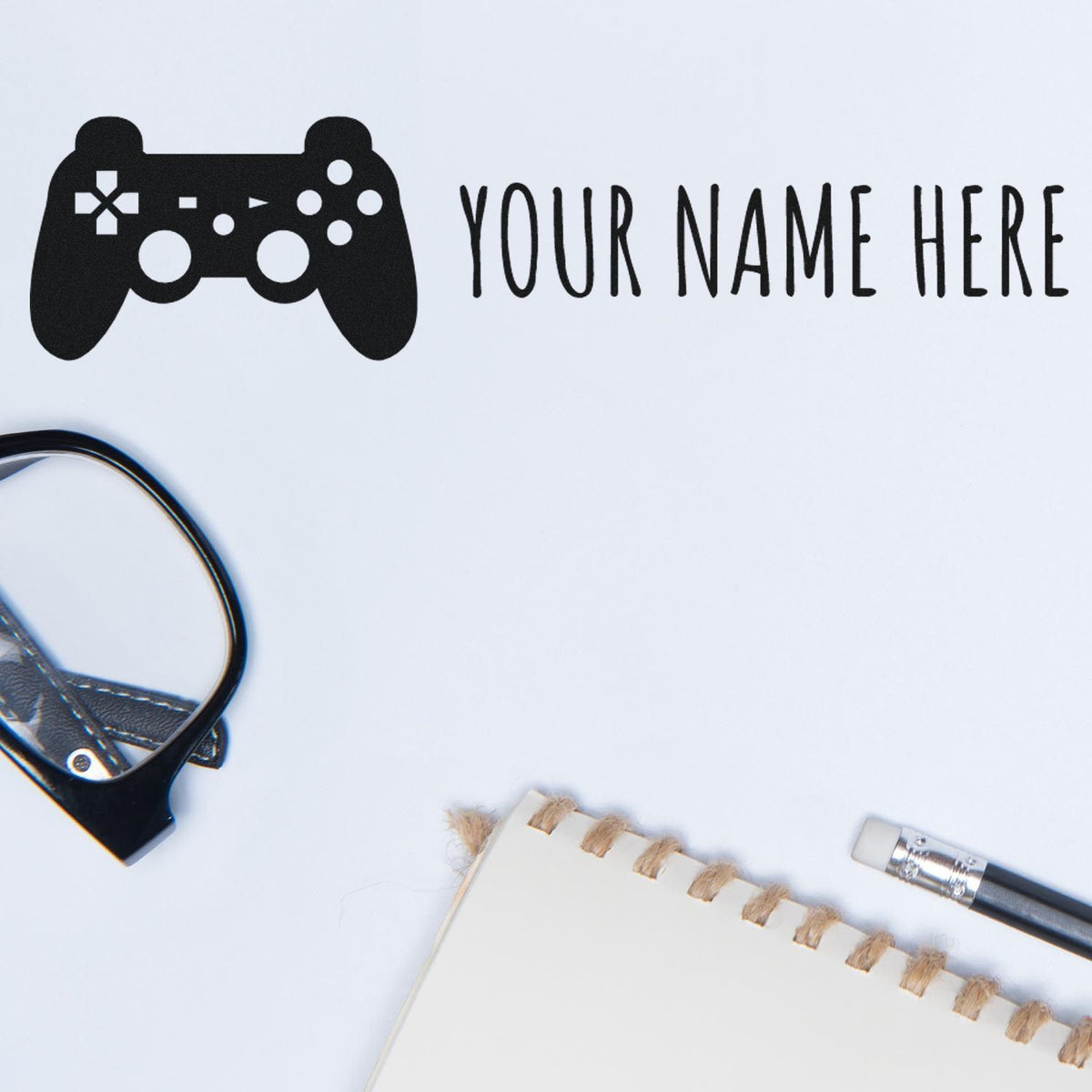Black custom game controller stamp and seal design with YOUR NAME HERE on white, beside notebook and pen.