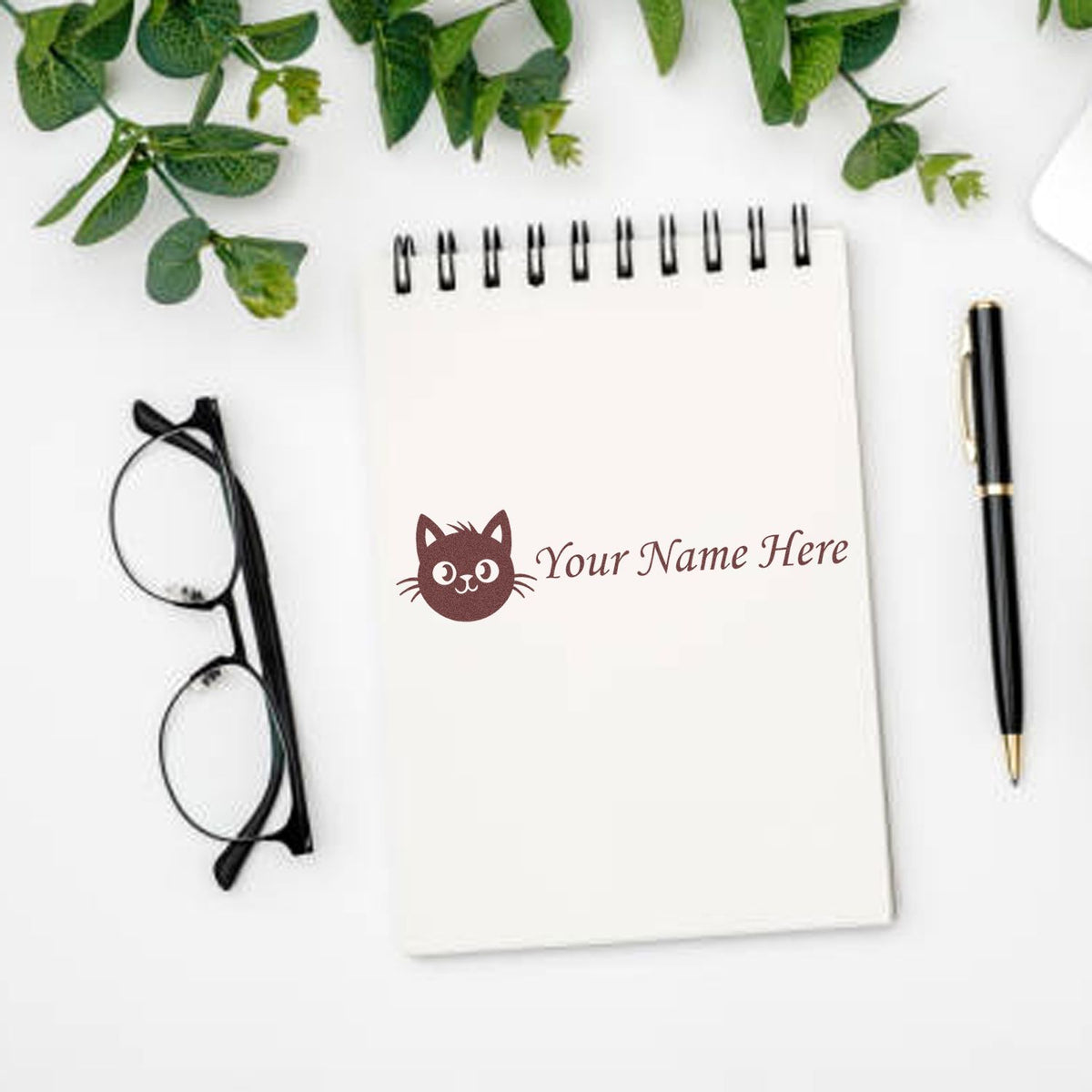 Custom cat name stamp in brown on a white spiral notebook, with black pen, glasses, and green leaves on a white desk.