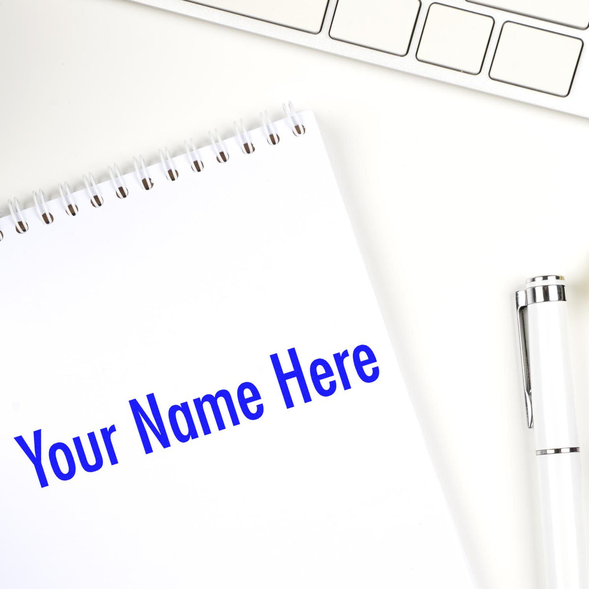 Custom stamp and seal on white spiral notebook with blue Your Name Here text, white pen, and keyboard.