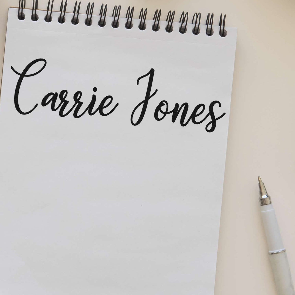 Custom black Carrie Jones stamp and seal on white spiral notepad with white pen on beige background.