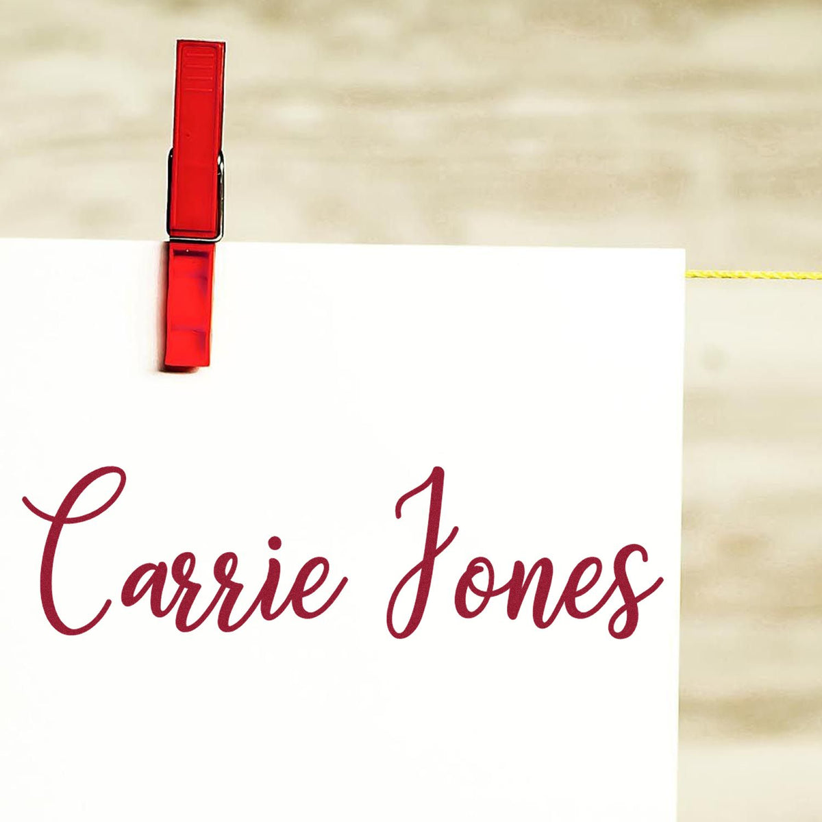 Red stamp-and-seal clip holding a white card with burgundy Carrie Jones lettering on a beige background.