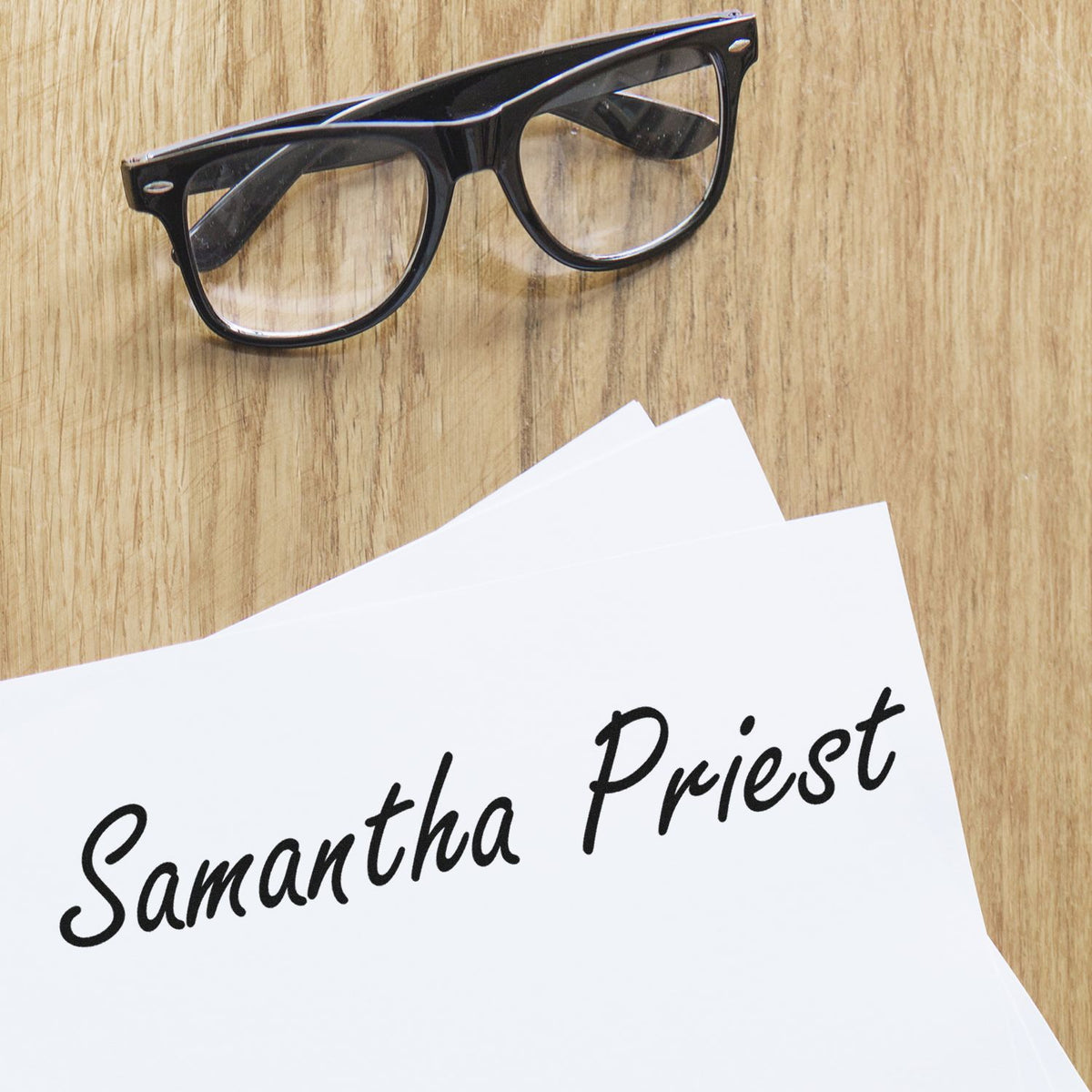 Black eyeglasses beside a white custom stamp and seal sheet reading Samantha Priest on a light wood background.