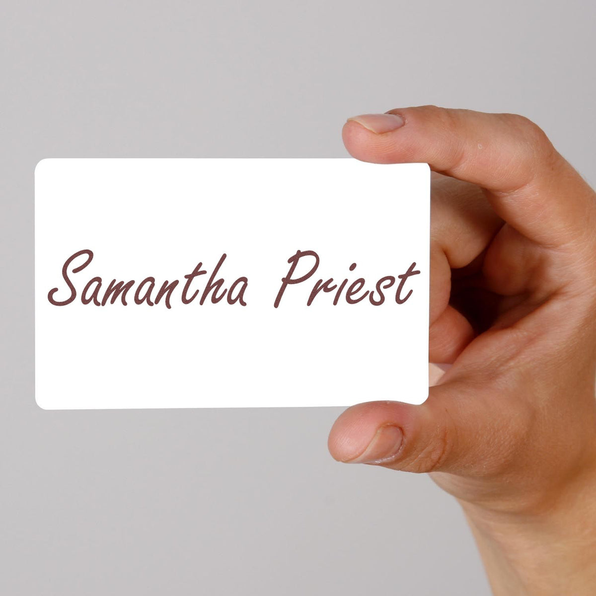 White stamp and seal card with brown script Samantha Priest held between fingers on a gray background.