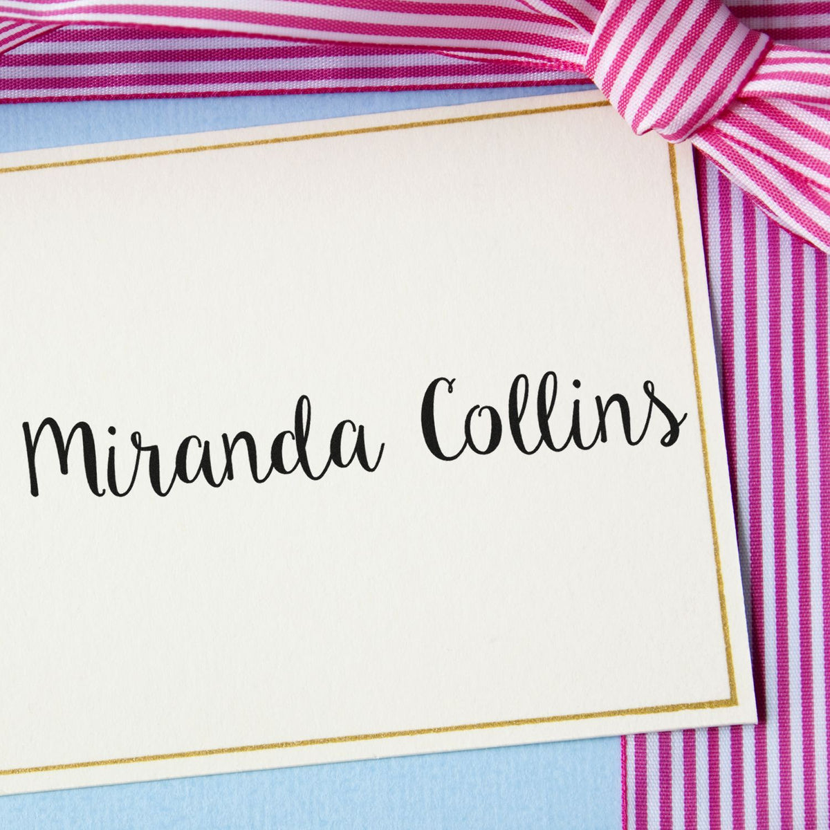 Custom stamp and seal for Miranda Collins, white card with gold border, black script, pink striped ribbon, and light blue background.