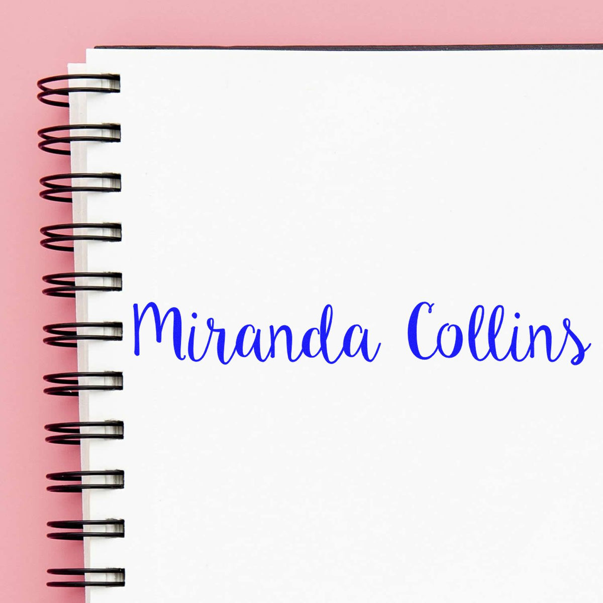 Blue Miranda Collins stamp and seal on a white spiral notebook with a pink background.