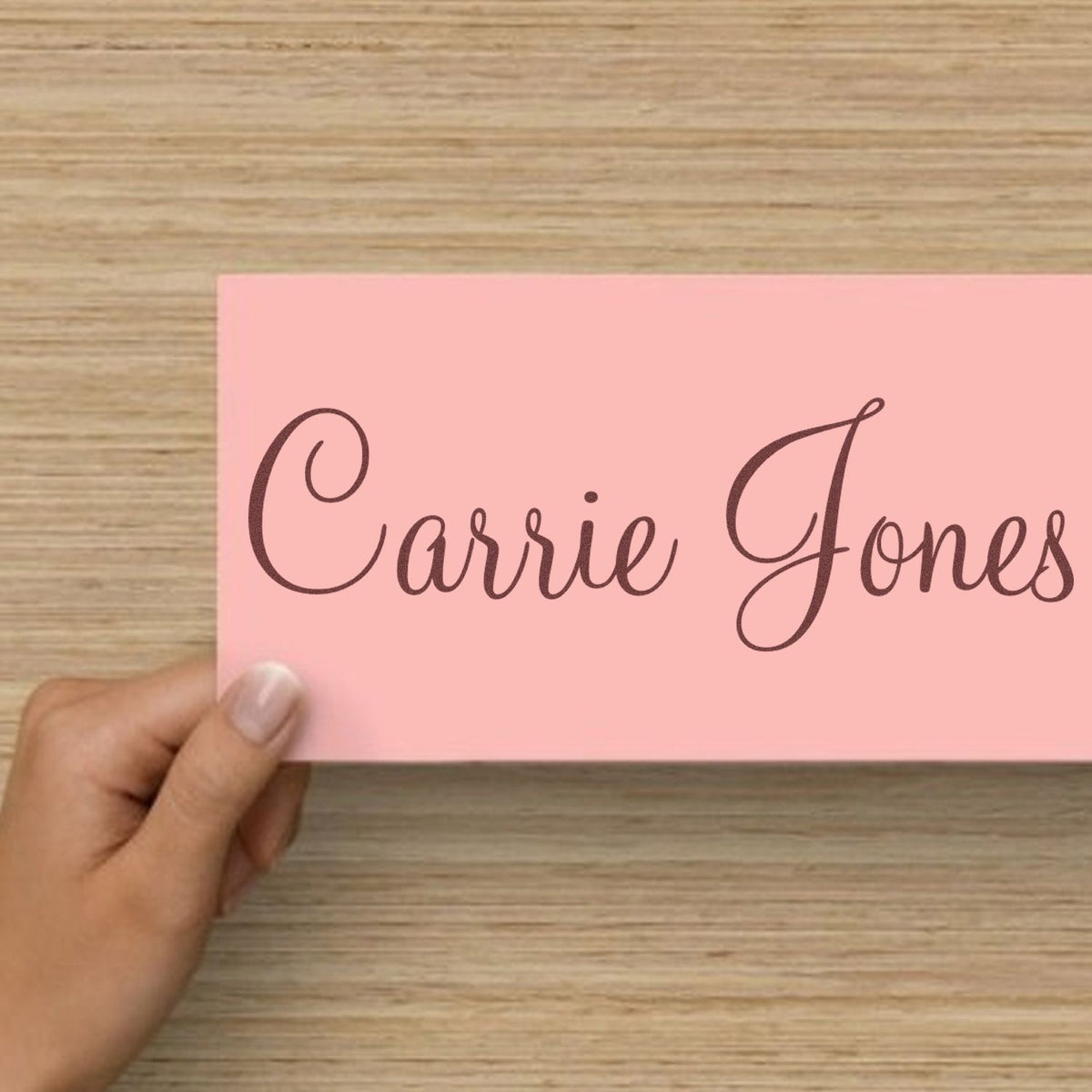 Pink custom stamp and seal card with dark brown cursive Carrie Jones held over a wood surface.