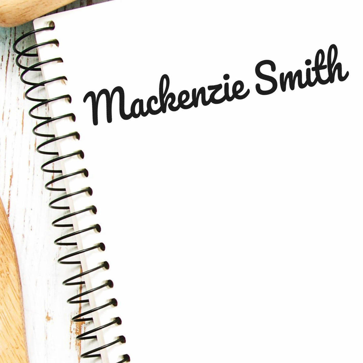 Black script name stamp reading Mackenzie Smith on a white spiral notebook with wood accents.