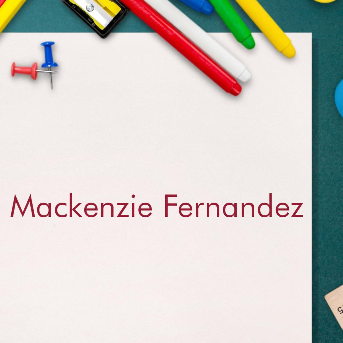 White stamp and seal card with Mackenzie Fernandez in red, surrounded by colorful markers and push pins on teal background.