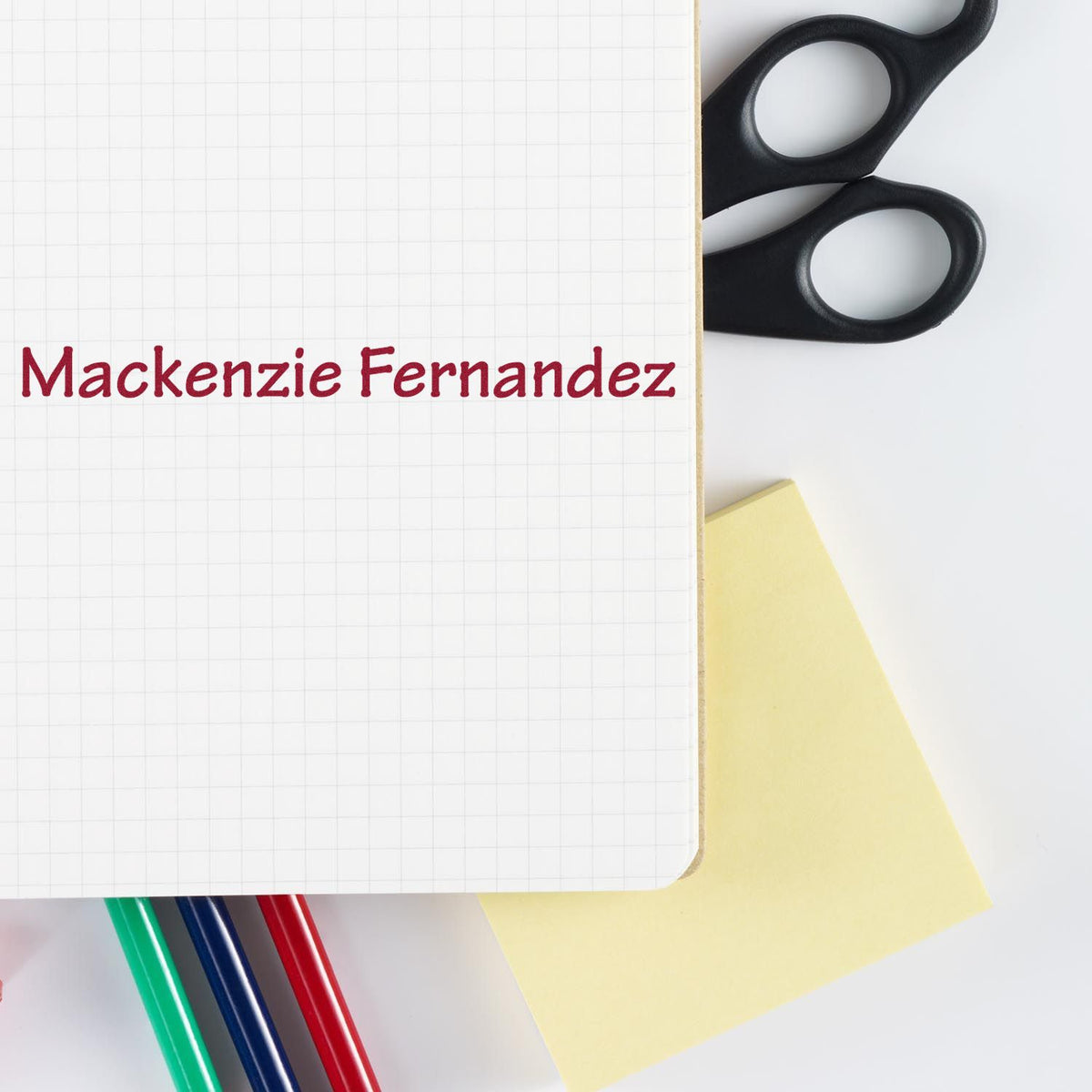 Custom red stamp and seal reading Mackenzie Fernandez on graph paper, with black scissors, yellow note, and colored pens.