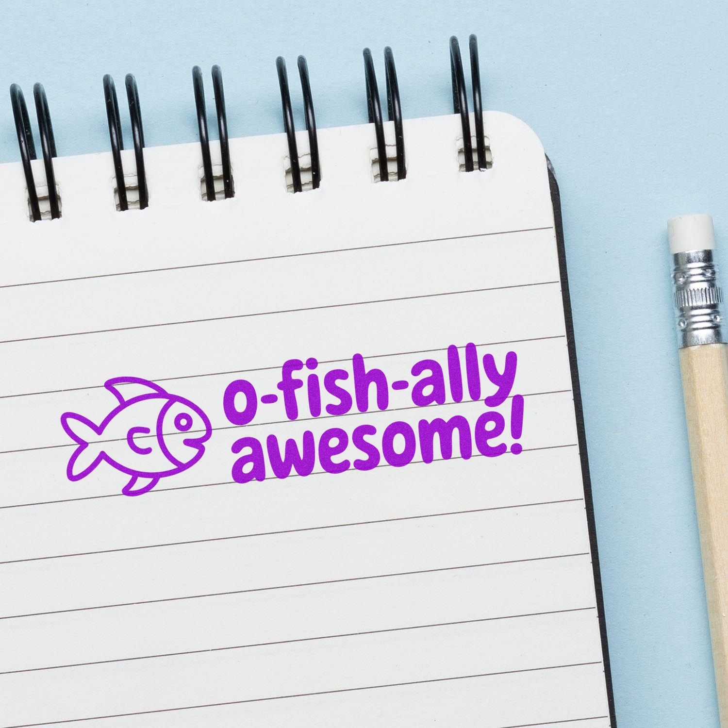 Purple fish stamp reading O-fish-ally awesome! on white spiral notepad with light blue background and pencil.