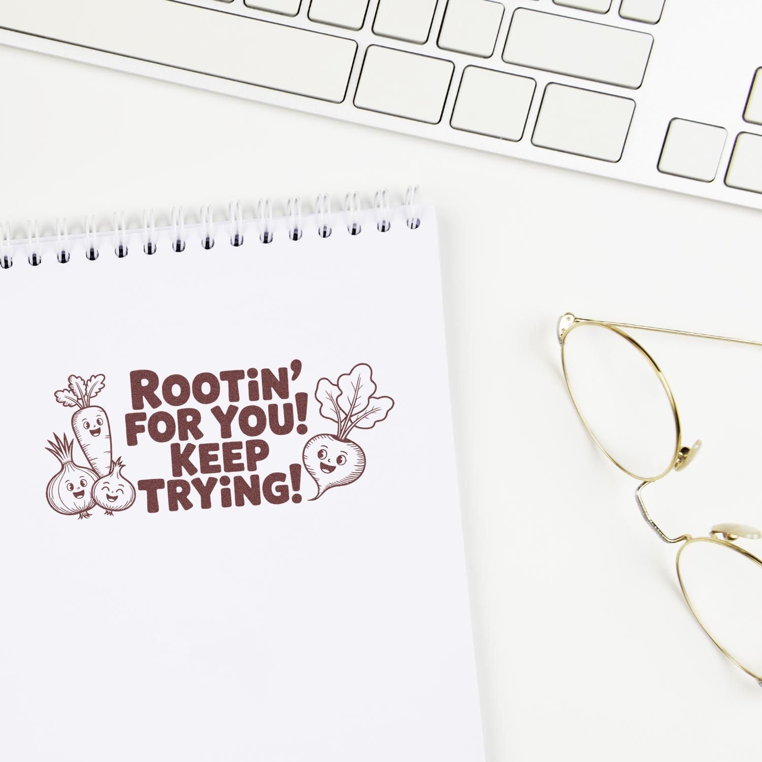 Brown vegetable stamp seal on notepad reads Rootin for you! Keep trying! beside keyboard and gold glasses.