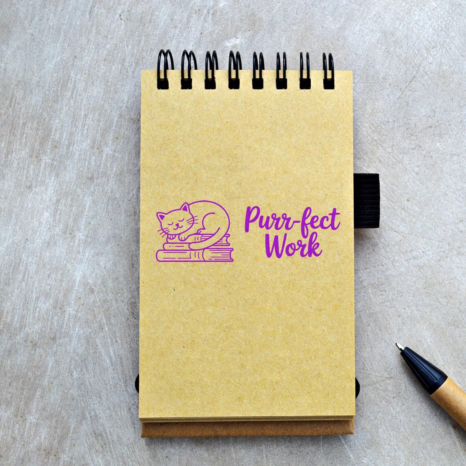 Yellow spiral notebook with purple cat Purr-fect Work stamp seal, black coil binding, and pen on gray desk.