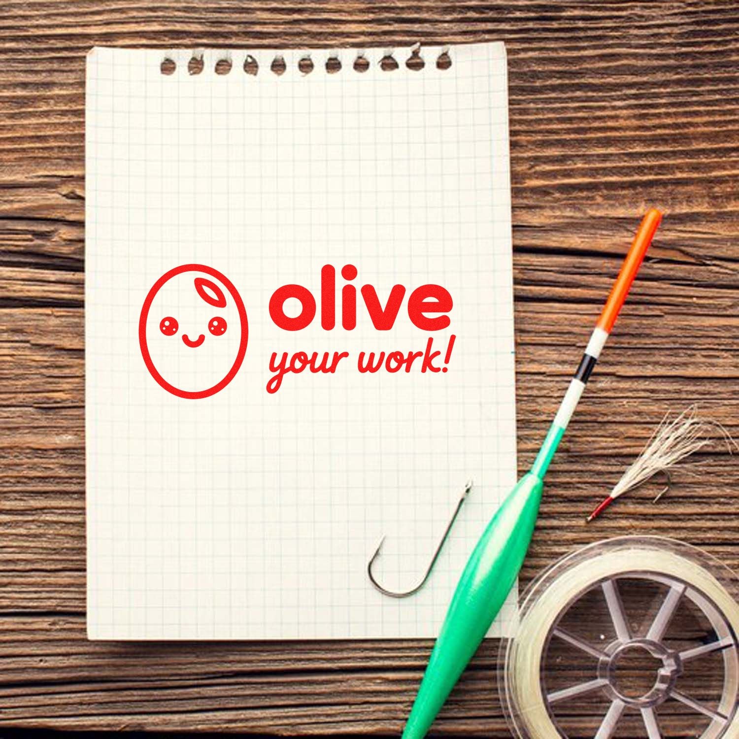 Red olive your work stamp with smiley face on graph notepad, beside green seal tool, orange pen and tape on wood table.