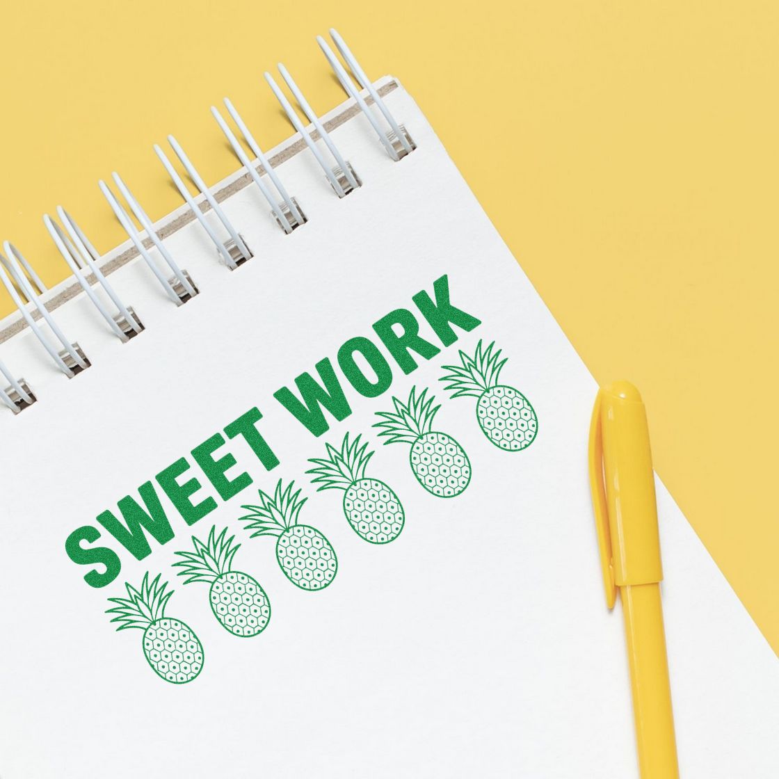 White spiral notebook with green Sweet Work pineapple stamp and seal design and yellow pen on a yellow background