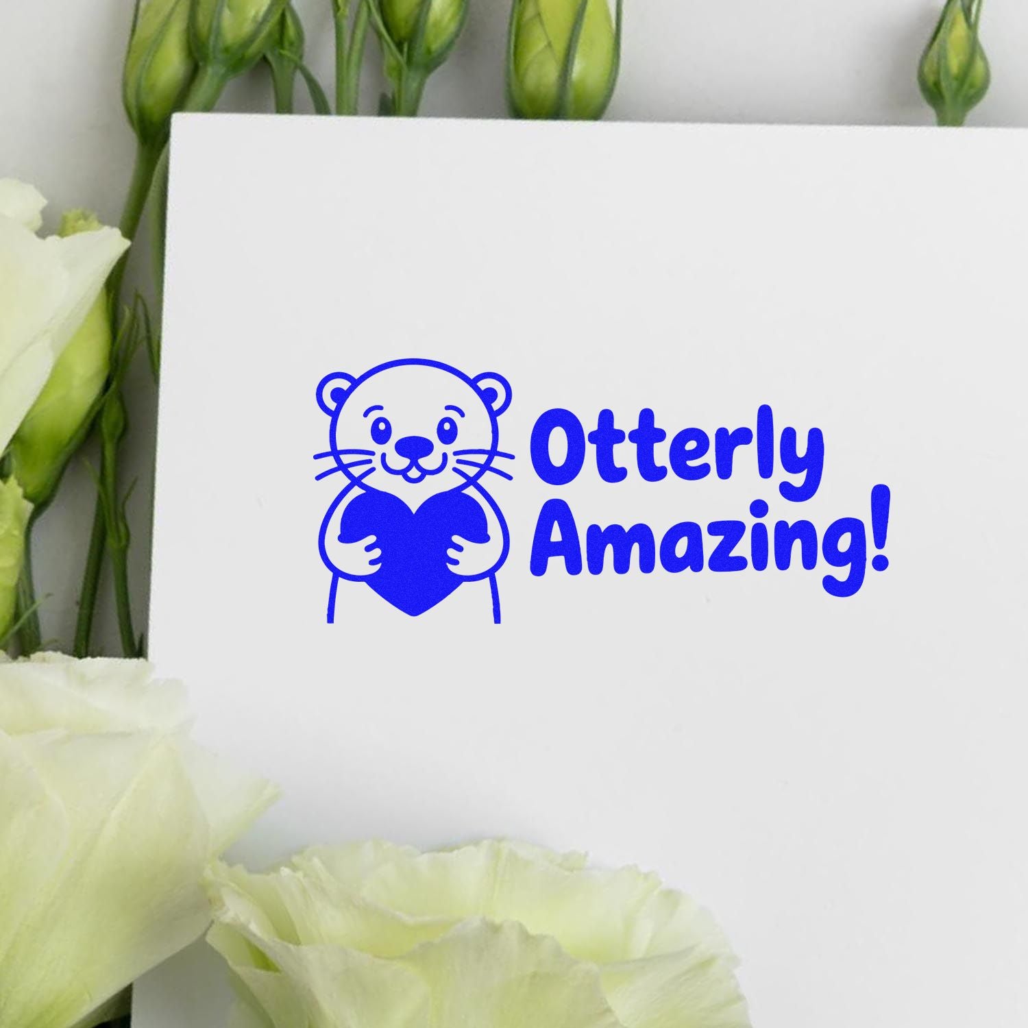 Blue otter stamp and seal design reading Otterly Amazing! on a white card, framed by pale white flowers.